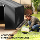 Man setting up grey 10x10 portable shed with ground stakes on a grassy area.