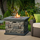 Outdoor stone propane fire pit with flames and a glass of beer on a patio with greenery.