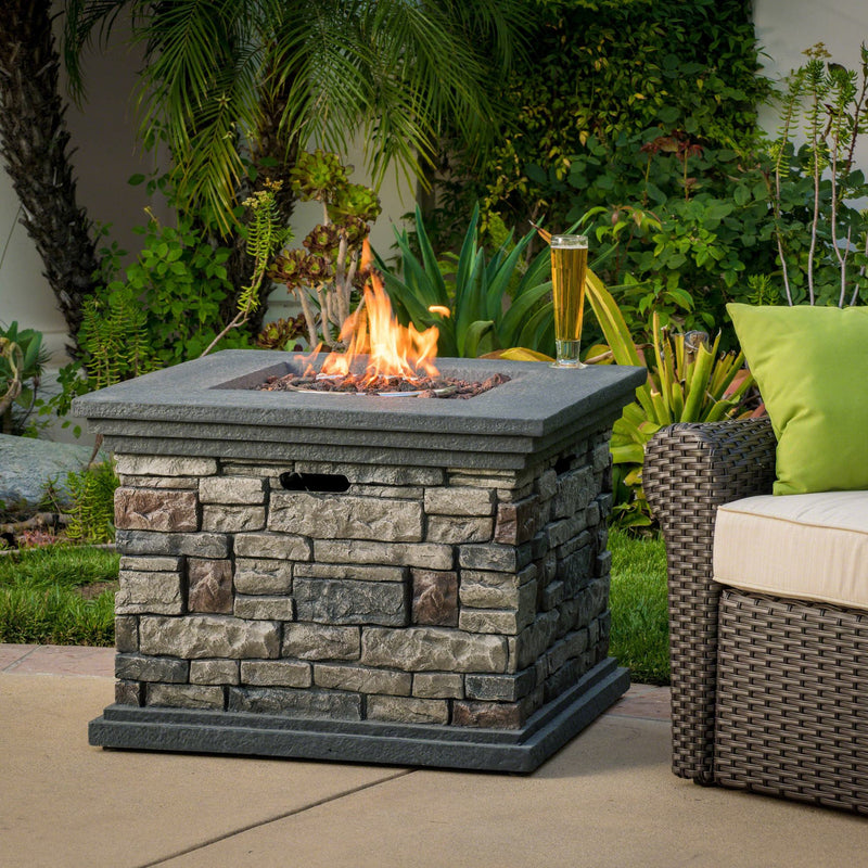 Outdoor stone propane fire pit with flames and a glass of beer on a patio with greenery.