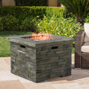 Stone-patterned MGO propane fire pit table with flames on a patio, surrounded by greenery and outdoor furniture.